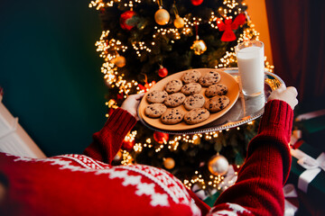 Festive moment as a person in a red sweater carries a silver tray of cookies and milk before a glowing Christmas tree during a cozy home holiday scene