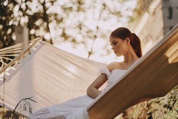 Young woman in white dress relaxing in a hammock, enjoying a serene moment in nature during golden hour, evoking feelings of peace and tranquility