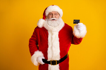 Santa Claus in a red suit with white fur trim holds a card against a bright yellow background for festive holiday shopping and celebration