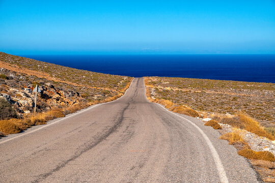 Road to the sea near Kato Zakros, Crete, Greece, showing empty asphalt road through dry hills and rocky landscape leading to blue Mediterranean coast