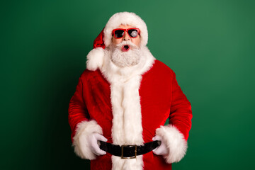 Santa in a red suit with white fur trim poses against a green background wearing a hat belt and bright red glasses with a playful expression