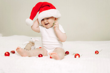Baby sitting on the bed wearing a Santa hat, surrounded by festive bokeh and Christmas ornaments. The child is exploring red and gold decorations with curiosity and joy.