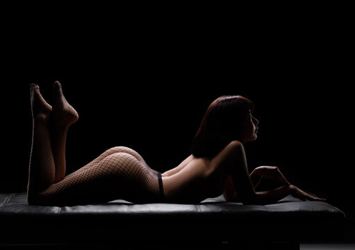 Artistic nude portrait of a woman in fishnet tights lying on a couch under dramatic backlight. Minimalist style, play of light and shadow, elegant body curves and sensual mood.