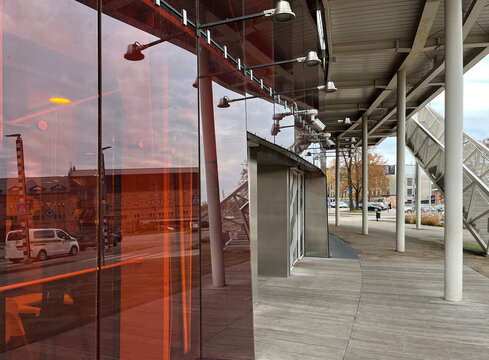 Modern architectural detail with red glass panels reflecting the surroundings, under a metal canopy with lights and structural beams, next to a paved urban area.