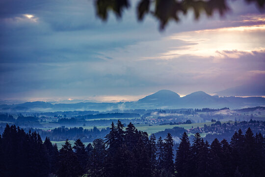 Misty sunrise over the Allgäu landscape with forest silhouettes and rolling hills under a dramatic cloudy sky in southern Germany