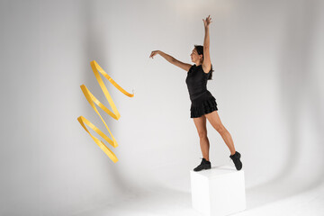 A graceful adult female rhythmic gymnast in a black costume and fishnet stockings performs a dynamic routine with a swirling yellow ribbon while balancing on a white cube against a plain studio 