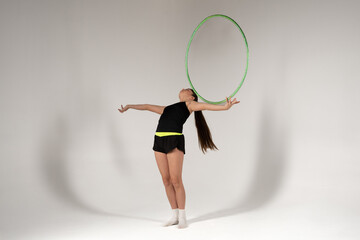 A flexible female performer with a long ponytail, dressed in sportswear, gracefully balances a green hula hoop on her face while leaning back with outstretched arms against a white studio background