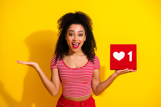 Smiling woman holding social media like icon box against a vibrant yellow background with an expressive and cheerful pose