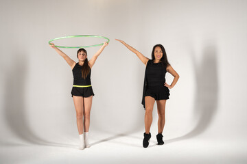 A young rhythmic gymnast holds a green hoop above her head while standing next to her smiling adult coach or partner, both posing in black athletic attire for a full-length portrait in a studio with a