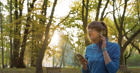 Young Caucasian woman with wired earphones listening music. Choosing best song or playlist on her smartphone. Preparing for running in beautiful park. Man jogging in background. Active lifestyle.