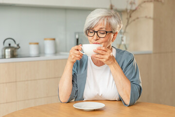 Good morning. Comfort relaxed senior old elderly woman grandmother drinking hot beverage tea coffee in kitchen at home. Mature woman resting enjoy calm morning at modern home alone. Pension concept