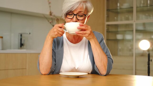 Good morning. Comfort relaxed senior old elderly woman grandmother drinking hot beverage tea coffee in kitchen at home. Mature woman resting enjoy calm morning at modern home alone. Pension concept