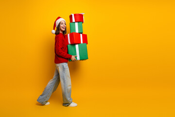 Funny santa girl carries stack of gifts in red sweater and jeans against bright yellow background...