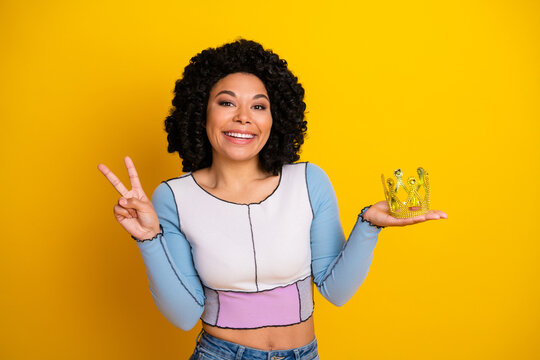Fototapeta Young woman with a cheerful expression holding a crown on yellow background, showcasing happiness and positivity