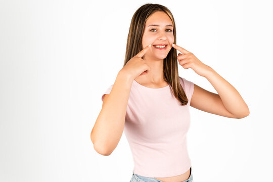 Teenage girl with straight brown hair in a pink shirt points to her smile, standing against an isolated white background. Suitable for concepts related to dental care, happiness, youth, and positive