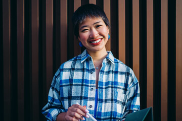 Cheerful Asian woman with short hair standing confidently with digital pen, portraying friendly attitude and creative energy, embodying casual professionalism in digital environment.