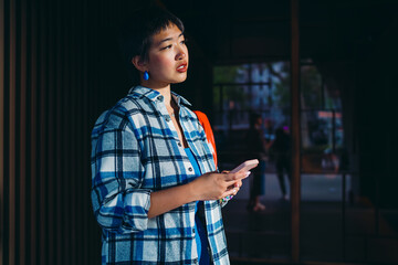 Asian woman with phone gazing into distance in afternoon light, conveying digital disconnection,...