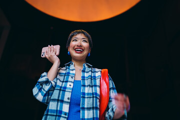 Asian woman laughing with smartphone raised mid‑gesture, symbolizing the joy of mobile voice/assistant tech, expressive digital moment and energetic mobile lifestyle.