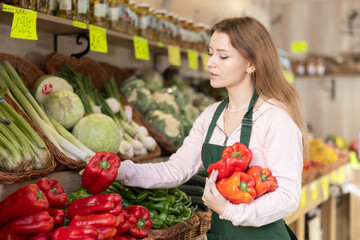 Young woman seller in apron puts fresh pepper on display at vegetable market