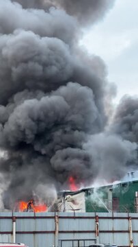 Massive fire engulfing an industrial building, sending thick plumes of black smoke into the sky as firefighters battle the raging blaze