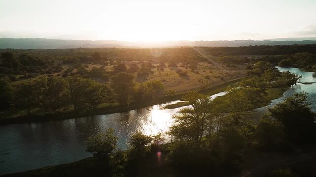 vista a&eacute;rea del atardecer la costa de un r&iacute;o con el sol y los arboles reflej&aacute;ndose en su espejo de agua y las monta&ntilde;as de fondo