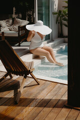Female relaxing by poolside in white swimsuit and wide-brim hat on sunny day