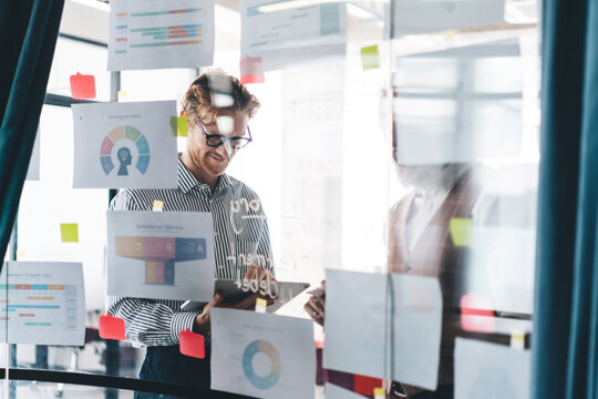 Two colleagues review printed charts on office glass wall, smiling as they discuss tech-enabled planning using tablet and infographics. Symbolizes data fluency and creative direction.