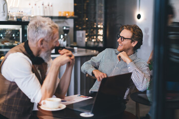 Freelancers laugh during break in coffee shop workspace, exchanging ideas in casual environment. Scene reflects startup ease, creative trust and digital-age team bonding.