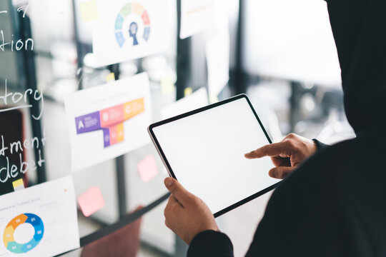 Person explores blank tablet screen beside transparent wall covered with strategy charts. Captures raw digital exploration, ideation stage, and early-phase tech project design.