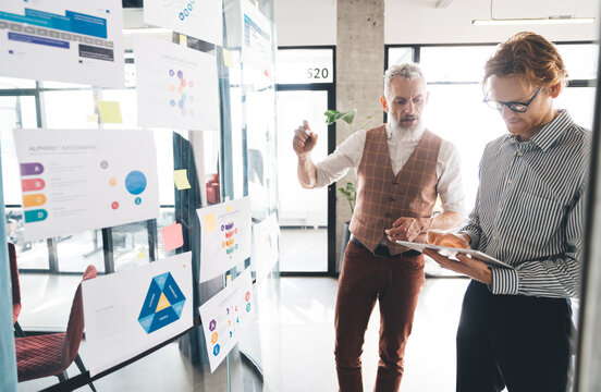 Senior businessman points at wall charts while explaining to younger colleague holding tablet. Image symbolizes active leadership, digital integration, and collaborative strategy process.