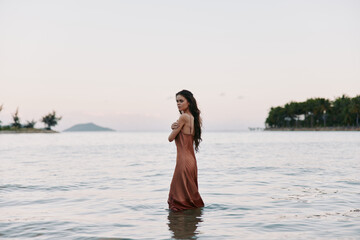 A solitary woman in a flowing bronze dress stands waist deep in calm sea, facing the shore as soft light bathes the scene in warm pastel tones.