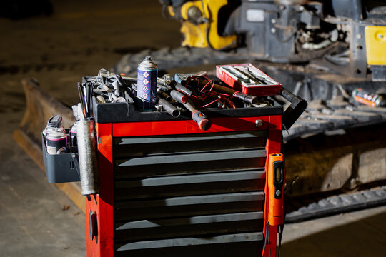 Red mechanic tool cabinet filled with hand tools, lubricants, and electrical cables placed beside construction machinery showing maintenance setup, workshop organization - Powered by Adobe