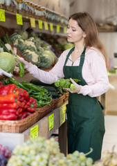 Young woman seller in apron puts fresh pepper on display at vegetable market