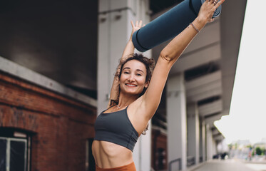 Fototapeta premium Smiling young woman lifting yoga mat overhead with joy and energy. Conceptual wellness photo symbolizing empowerment, mindfulness, individuality, and digital-era connected lifestyle culture.