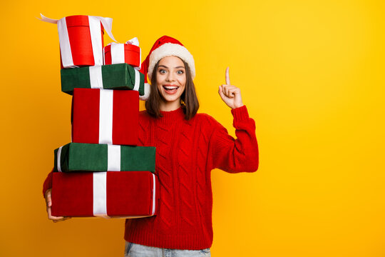 Playful woman in a Santa hat carries a towering stack of gifts against a bright yellow background creating a festive and family friendly holiday moment