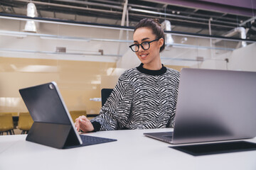 Smiling freelancer interacting with tablet and laptop at clean white desk, symbolizing digital collaboration, productivity and joyful remote work culture in modern creative office.