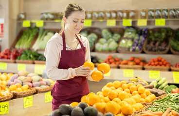 Obraz premium Young woman seller in apron puts fresh oranges on display in vegetable shop