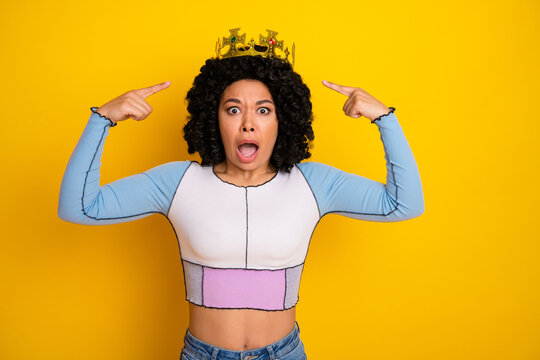 Fototapeta Confident young woman points to crown with surprised expression, standing against vibrant yellow background wearing casual fashion