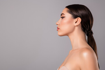 Elegant profile portrait of a young woman with flawless makeup and sleek hairstyle posing against a grey background.