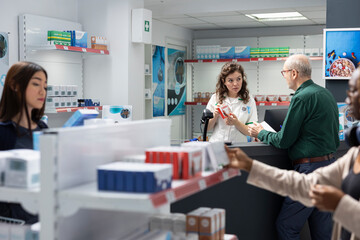 Old male client speaking with a pharmacy worker at the drugstore checkout, seeking advice on prescription drugs, over the counter pills and healthcare products available on shelves.