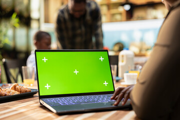 Green screen laptop next to parents and kids sharing breakfast in the kitchen, serving eggs, croissants and orange juice. Bright and cozy domestic moment celebrates family bonding together.