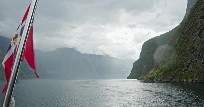 Norway fjord cruise with flag in foreground and dramatic cliffs near Flam on an overcast day, boat wake across calm water in a scenic mountain landscape