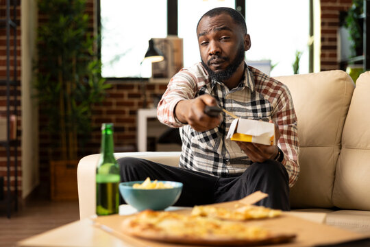 Young african american man seated on couch squinting at TV while flipping channels. Black guy holds remote control and takeaway food box in relaxed living room, enjoying casual meal at home. - Powered by Adobe