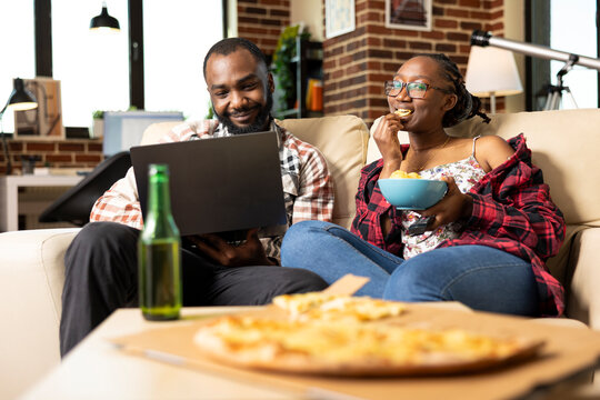 Happy african american couple relaxing on cozy sofa, enjoying leisure time at home. Bearded man works on laptop as girlfriend watches TV and eats snacks during peaceful weekend evening. - Powered by Adobe