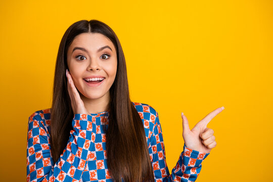 Bright surprised young woman with long brunette hair wearing a colorful top points to the side against a vivid yellow background