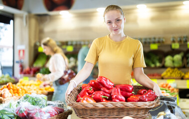 Teenage girl seller in apron puts fresh pepper on display at vegetable market