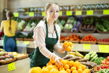 Young woman seller in apron puts fresh tangerines on display at vegetable market