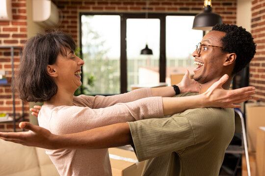 Cheerful young multiethnic couple hugging in living room during moving day. Smiling African American man and Caucasian woman celebrate home ownership with joy, bonding and affection.