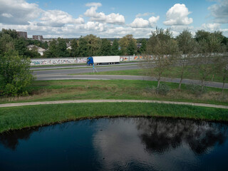 Fototapeta premium Blue truck with white trailer traveling on highway past water canal trees and residential buildings