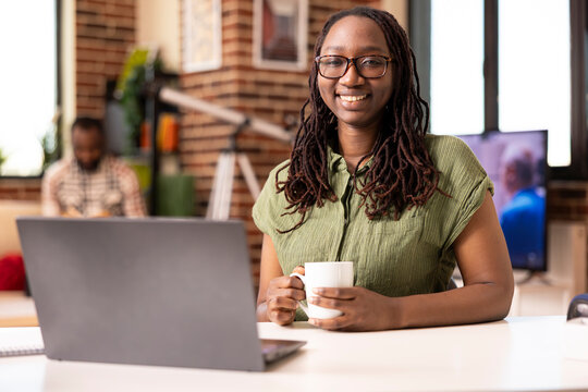 Portrait of smiling black female freelancer sitting at desk with laptop and coffee cup, taking break from remote work. Confident african american woman posing in casual home office setting.